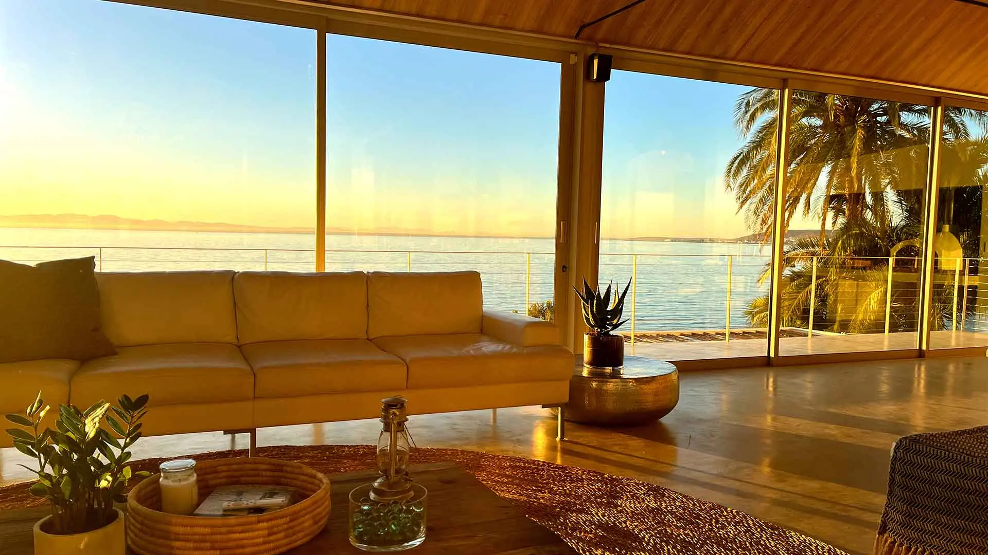 Golden sunrise light flooding the lounge, white sofa, palms and the bay beyond the glass