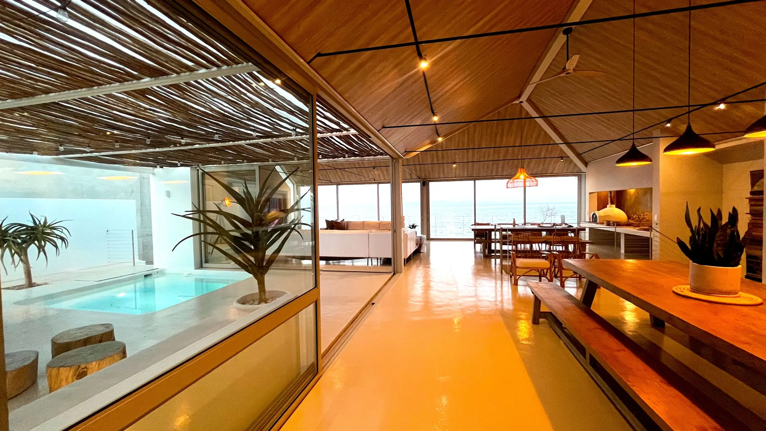 Looking along the living floor at dusk: harvest table, pendants, the lit courtyard pool through the glass